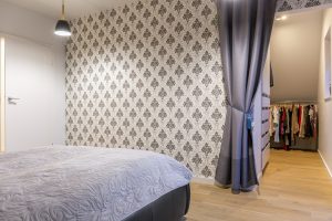 Modern bedroom with monochrome wallpaper, minimal bed, light wood flooring, and a walk-in wardrobe.