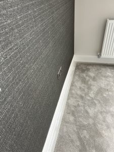 Modern living room corner with textured wallpaper, electrical outlet, plush carpet, skirting, and radiator.
