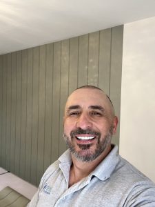 Smiling man in grey polo shirt taking a selfie in a stylish room with green wall panelling.
